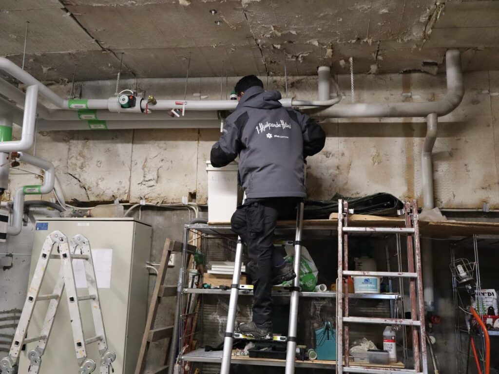 Vaktmester arbeider på en stige ved rør og tekniske installasjoner i et teknisk rom