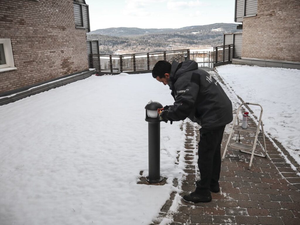 Vaktmester skifter lyskilde på en utelampe i et snødekt uteområde