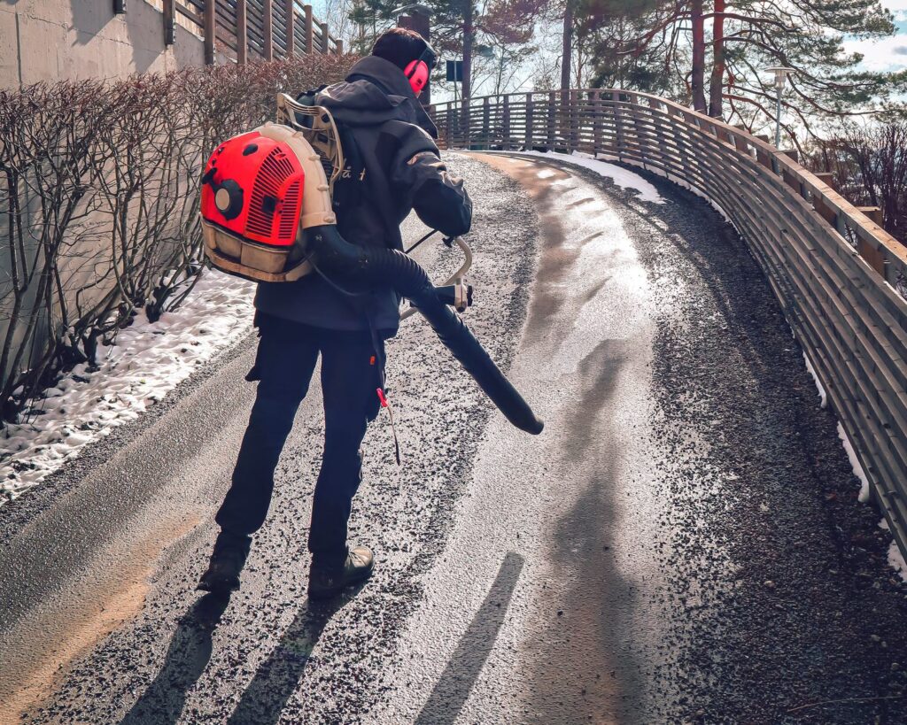 Vaktmester med løvblåser fjerner snø og strøsand fra en isete gangvei