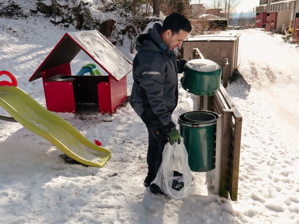 Vaktmester tømmer en avfallsdunk ved en snødekt lekeplass